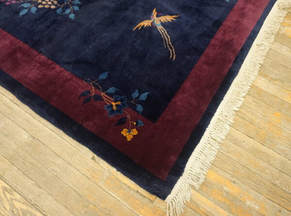 Navy blue rug with stylized gold bird and blue flowers bordered by maroon on a light wood floor.