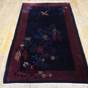 Navy blue rug with a floral vine pattern and bird on a maroon border sits on distressed wood floor.
