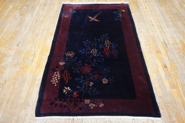 Navy blue rug with a floral vine pattern and bird on a maroon border sits on distressed wood floor.