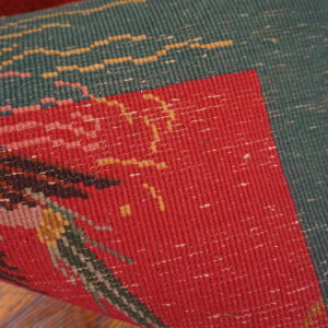 Tightly woven red and green rug, displaying a complex pattern, rolled up on brown wood flooring.