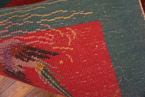 Tightly woven red and green rug, displaying a complex pattern, rolled up on brown wood flooring.