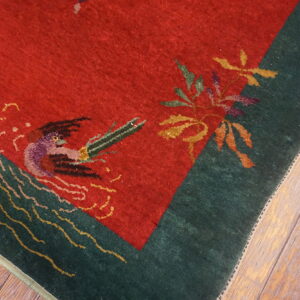 Red rug with a green border and detailed bird and leaf patterns on distressed wood flooring.