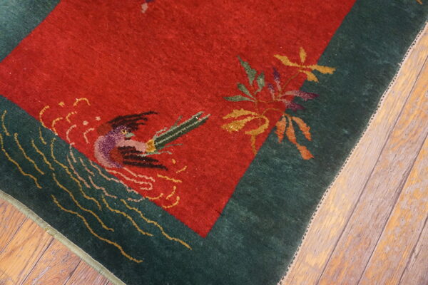 Red rug with a green border and detailed bird and leaf patterns on distressed wood flooring.