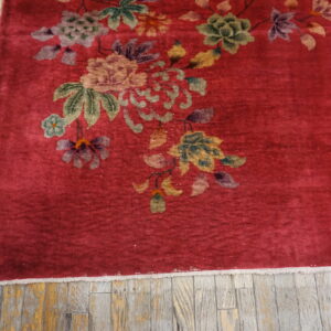 Crimson rug featuring multicolor floral patterns, bordered by fringe and set against distressed wooden planks.