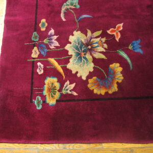 Burgundy plush rug displaying a corner floral motif in teal, gold, and blue on distressed wooden flooring.
