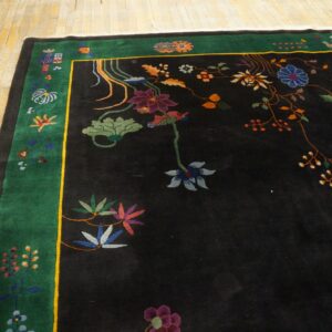 A black rug with a high pile features multicolored floral designs and a wide green border on distressed yellow wood floorboards.