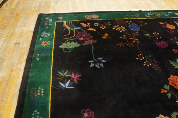 A black rug with a high pile features multicolored floral designs and a wide green border on distressed yellow wood floorboards.