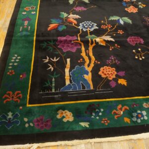 Black rug with colorful flowers and trees, bordered in green and gold, on light wood flooring.