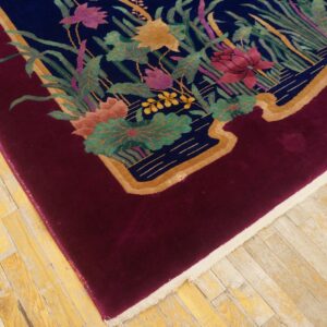 Plush rug corner featuring a deep red border and central multicolored floral design on light wood flooring.