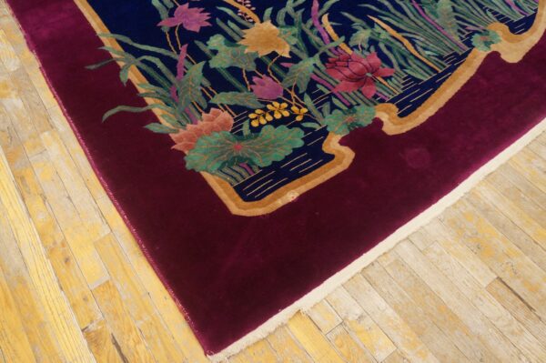 Plush rug corner featuring a deep red border and central multicolored floral design on light wood flooring.