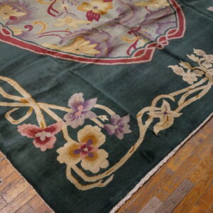 Corner of a deep green rug featuring ivory, pink, and purple floral border resting on distressed wood flooring.