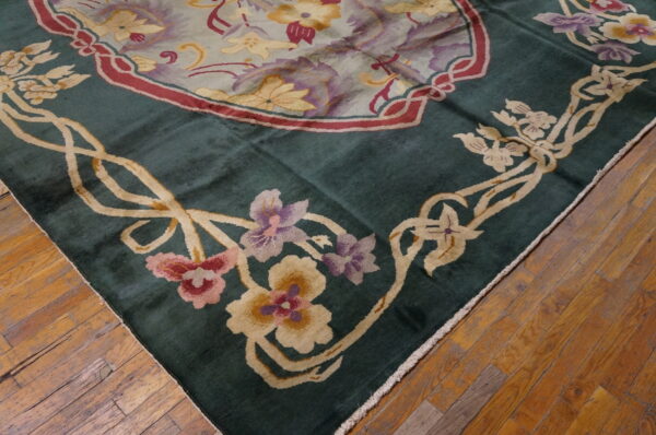 Corner of a deep green rug featuring ivory, pink, and purple floral border resting on distressed wood flooring.