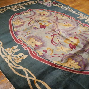 Teal rug with a gold floral border surrounding a central medallion of purple, gold, and burgundy flowers on wood flooring.