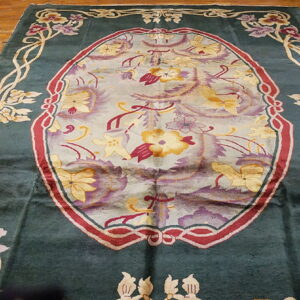 Dark green floral rug featuring a central light blue oval medallion and gold vine borders on wood flooring.