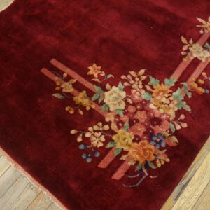Plush crimson rug with a multicolored floral and ribbon design resting on light wood floorboards.