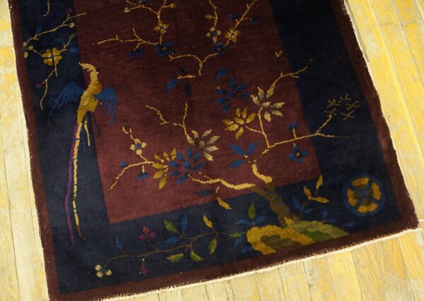A low pile navy and maroon pictorial rug depicting a gold bird and flowering branches on distressed wood flooring.