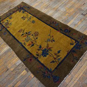 Gold field rug with a brown border and dark blue floral vines laid upon distressed wood flooring.
