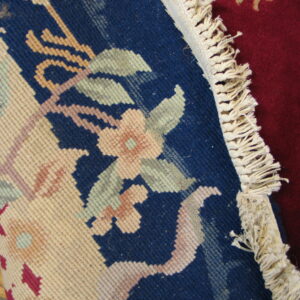 Rolled blue rug with floral pattern, cream fringe, and deep red rug visible on a wooden floor.