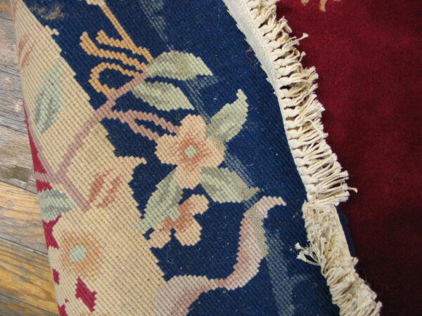 Rolled blue rug with floral pattern, cream fringe, and deep red rug visible on a wooden floor.