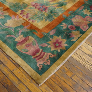 Gold and teal low-pile rug with complex floral motifs and a rust border on worn plank wood flooring.