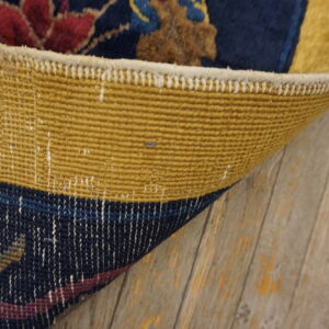 Rug corner flipped showing the yellow woven backing contrasting with the dark blue pile and distressed wood floor.