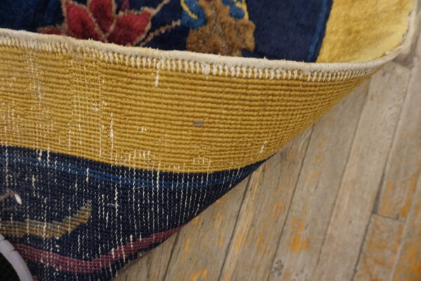 Rug corner flipped showing the yellow woven backing contrasting with the dark blue pile and distressed wood floor.