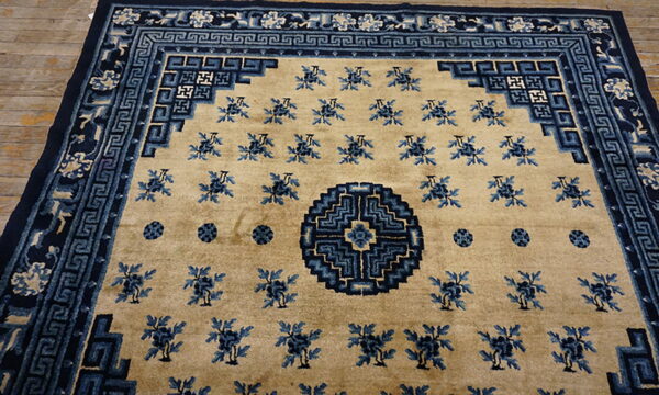 Tan rug with dark blue geometric borders, scattered florals, and a stepped circular central motif.