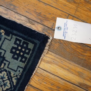 A navy and teal geometric rug corner with fringe rests on rustic wooden planks next to a product tag.