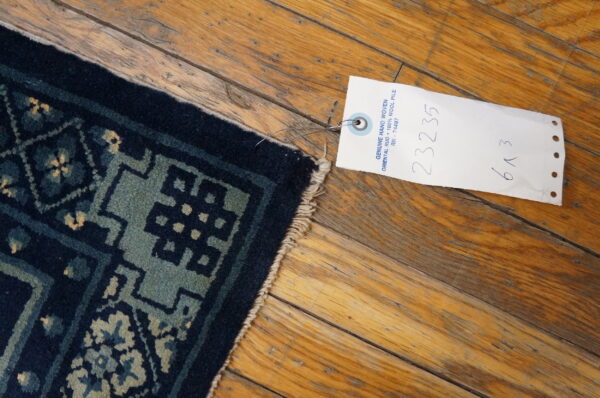 A navy and teal geometric rug corner with fringe rests on rustic wooden planks next to a product tag.