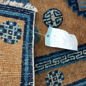 Close-up of a textured tan and blue rug showing geometric patterns, fringe, and an attached inventory tag.