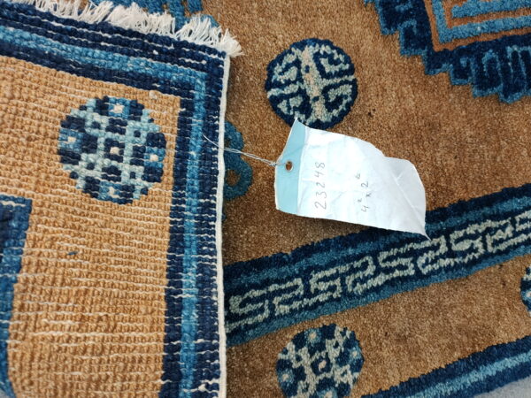 Close-up of a textured tan and blue rug showing geometric patterns, fringe, and an attached inventory tag.