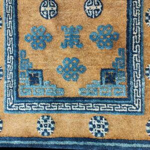 Textured tan rug with dark blue borders framing stylized blue floral patterns and white meander motifs.