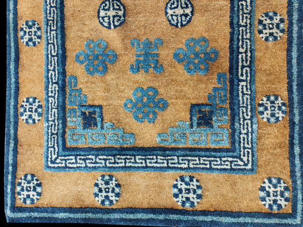 Textured tan rug with dark blue borders framing stylized blue floral patterns and white meander motifs.