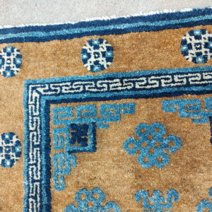 Tan rug with textured pile features blue meander borders and stylized circular floral motifs on gray pavement.