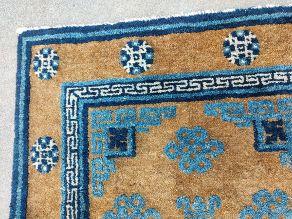 Tan rug with textured pile features blue meander borders and stylized circular floral motifs on gray pavement.
