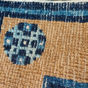 Textured rug close-up showing tan loops, a blue striped border, and a geometric circle pattern.