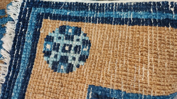 Textured rug close-up showing tan loops, a blue striped border, and a geometric circle pattern.
