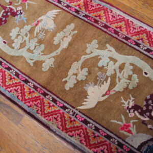 Close-up of a tan rug with pale blue branches and birds, bordered by red geometric patterns, lying on wood floor.