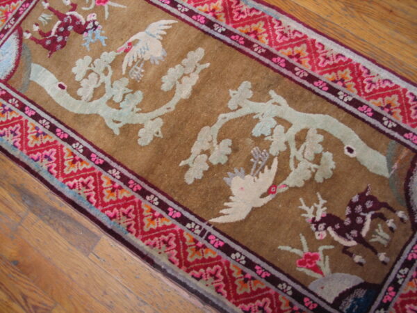 Close-up of a tan rug with pale blue branches and birds, bordered by red geometric patterns, lying on wood floor.