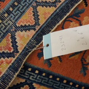Detailed view of a rust and navy rug border and field, including a white tag with handwritten numbers.