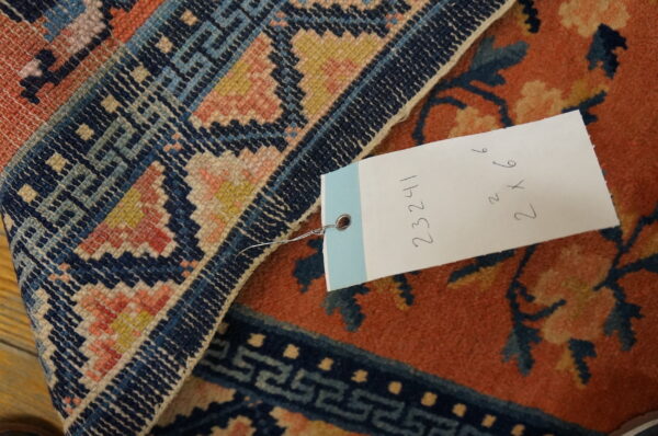 Detailed view of a rust and navy rug border and field, including a white tag with handwritten numbers.