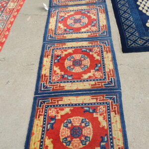A red, blue, and gold runner with geometric squares is displayed on gray concrete next to two other rugs.