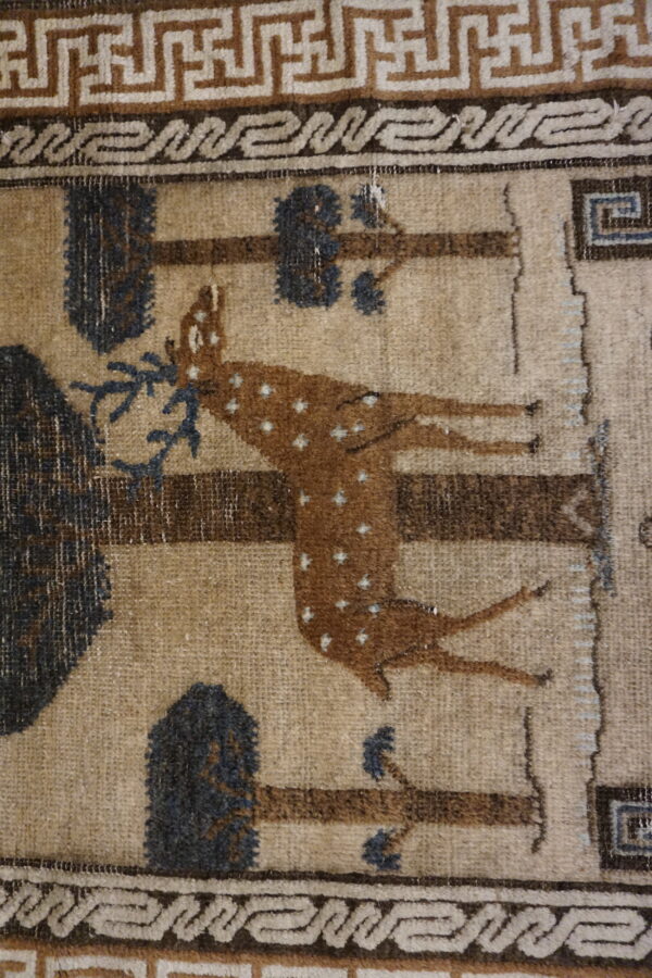 Tan and brown patterned rug with a spotted deer motif, bordered by meander and scroll designs.