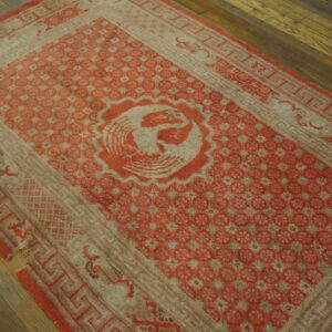 Worn red and pale beige rug featuring a central bird motif and geometric borders rests on dark wood flooring.