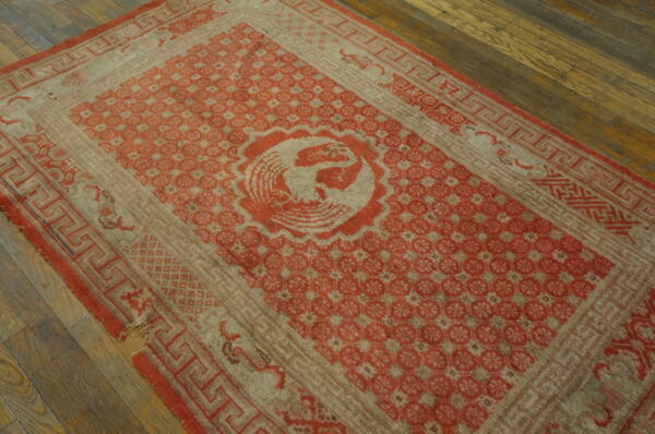 Worn red and pale beige rug featuring a central bird motif and geometric borders rests on dark wood flooring.