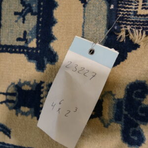A close-up view of a cream and blue patterned rug with an attached white inventory tag displaying numbers.