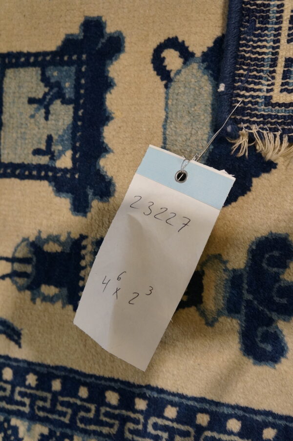 A close-up view of a cream and blue patterned rug with an attached white inventory tag displaying numbers.