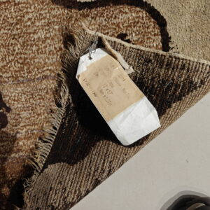 Close-up of a brown and tan patterned rug with visible pile, fringes, backing, and attached tag on a light floor.