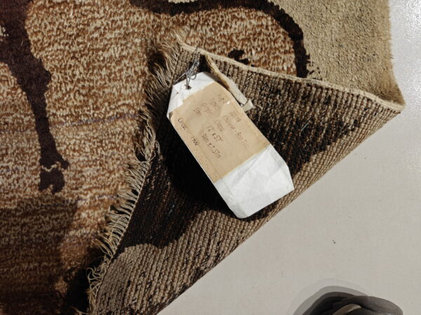 Close-up of a brown and tan patterned rug with visible pile, fringes, backing, and attached tag on a light floor.