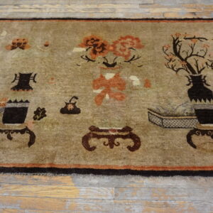 Muted tan rug with dark brown borders and stylized antique object patterns resting on worn wood flooring.